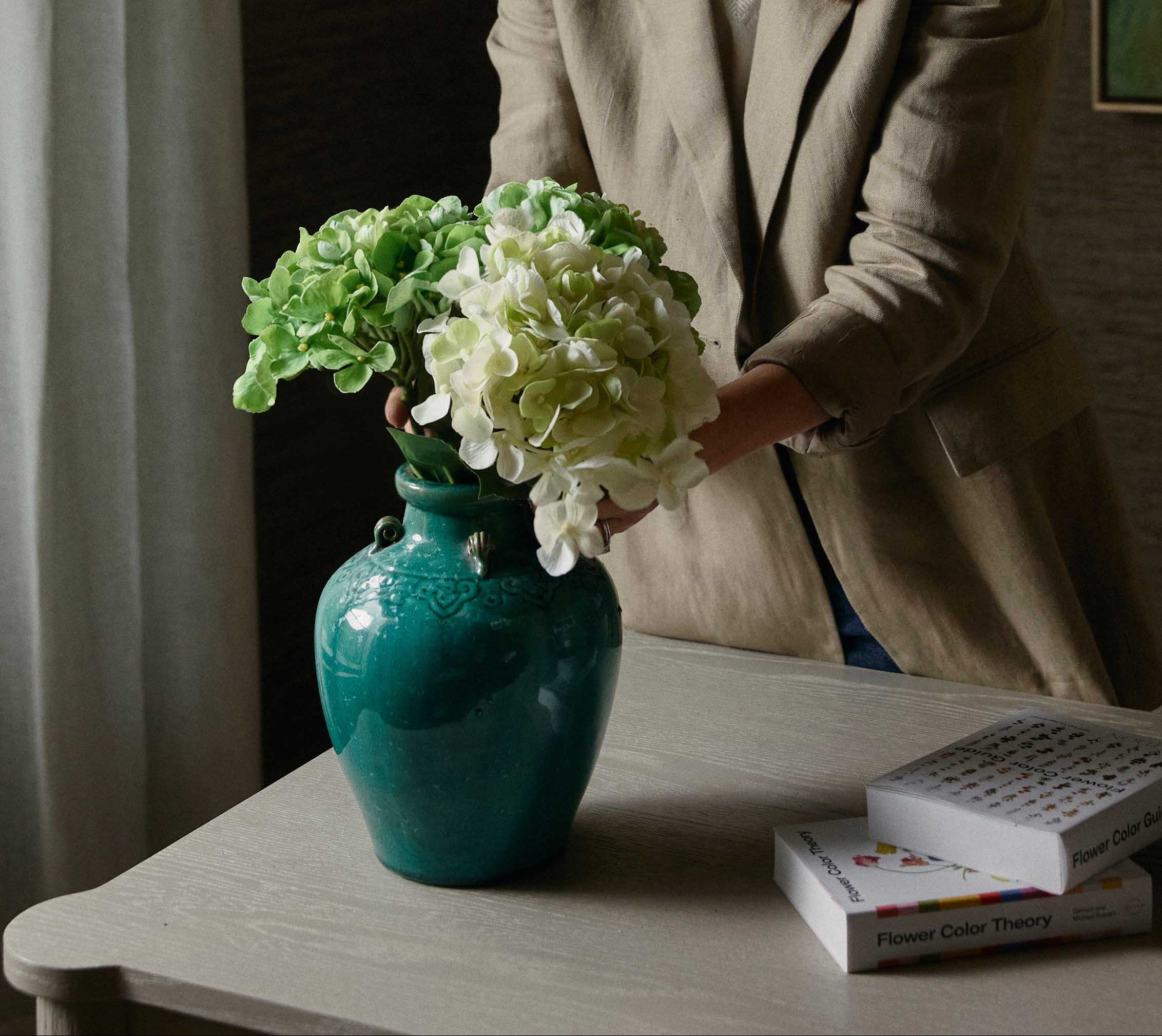 Woman standing in a room with a vase of artificial flowers and books on a table.